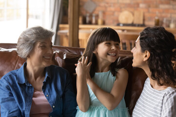 Close up overjoyed little girl with happy young mother and mature grandmother having fun, laughing, chatting, sitting on couch at home, three generations of women spending leisure time together