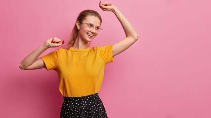Positive millennial girl dances carefree against pink studio background has upbeat mood wears casual yellow t shirt and skirt poses against pink background with copy space for your information