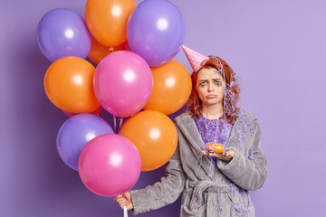 Fototapeta na wymiar Waist up shot of unhappy ginger woman frowns face and looks dissatisfied at camera has bad mood after birthday celebration holds cupcake with burning candle and inflated multicolored balloons