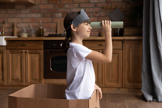 Side View Little Girl Playing Funny Game, Pretending Pirate, Adorable Child Kid Wearing Handmade Costume Holding Cardboard Tube As Telescope, Sitting In Box, Having Fun In Kitchen At Home