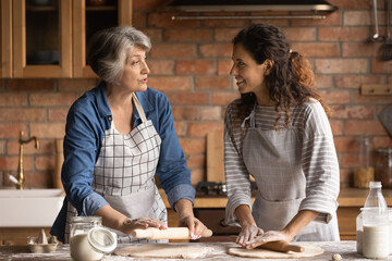 Mature grey haired woman with grownup daughter chatting, cooking homemade pastry, rolling dough,...