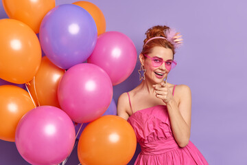 Fototapeta na wymiar Positive redhead woman indicates at you invites for party and celebration wears trendy sunglasses and dress holds bunch of inflated balloons celebrates anniversary isolated over purple background
