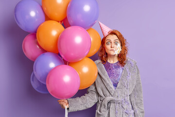 Fototapeta na wymiar Shocked pretty redhead woman has cake on face celebrates graduation from university enjoys free time on party poses with multicolored balloons wears dressing gown and paper cone hat on head.