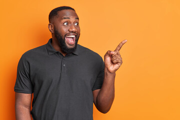 Happy amazed dark skinned man points at upper right corner demonstrates awesome advertismenet...