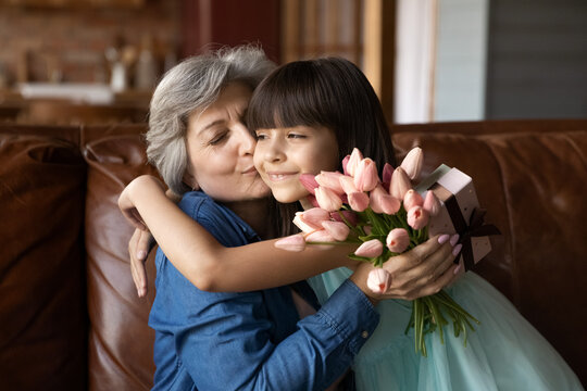 Close Up Little Granddaughter Congratulating Mature Grandmother, Hugging, Presenting Gift And Flowers, Thankful Older Woman Kissing Little Girl On Cheek, Family Celebrating Birthday Or 8 March