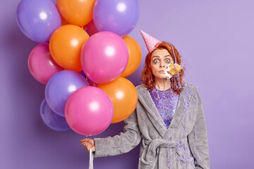 Fototapeta na wymiar Surprised birthday girl gets cake in face smeared with serpentine spray doesnt expect to get such congratulations from friends holds colorful balloons isolated over purple background. Holidays concept