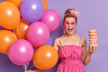 Positive redhead woman in trendy pink sunglasses and dress exclaims gladfully holds donuts has fun...