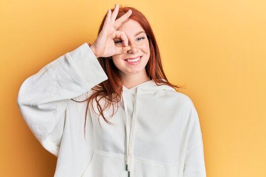 Young red head girl wearing casual sweatshirt doing ok gesture with hand smiling, eye looking through fingers with happy face.