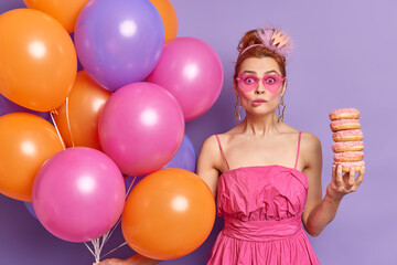 Photo of surprised female bites lips looks at camera through pink sunglasses poses with doughnuts and helium balloons dressed in festive clothes has fun on corporate party isolated over purple wall