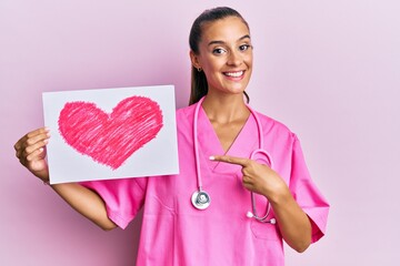 Young hispanic woman wearing doctor uniform holding heart draw smiling happy pointing with hand and...