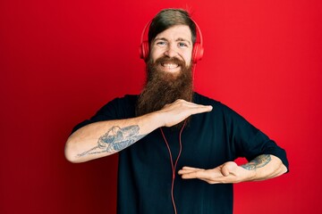 Redhead man with long beard listening to music using headphones gesturing with hands showing big and large size sign, measure symbol. smiling looking at the camera. measuring concept.
