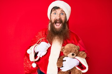 Handsome young red head man with long beard wearing santa claus costume holding teddy bear celebrating crazy and amazed for success with open eyes screaming excited.