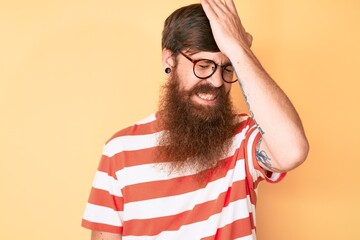 Handsome young red head man with long beard wearing casual clothes and glasses surprised with hand...