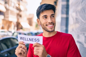 Young latin man smiling happy holding wellness word paper standing at the city.