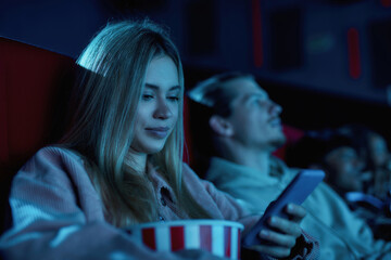 Portrait of beautiful young woman smiling, using her phone while missing boring movie at the cinema