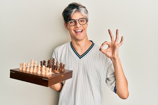 Young hispanic man holding chess doing ok sign with fingers, smiling friendly gesturing excellent symbol