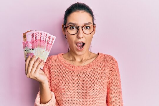 Young caucasian woman holding 100000 indonesian rupiah scared and amazed with open mouth for surprise, disbelief face