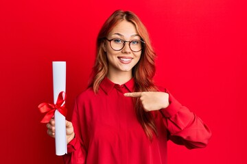 Young redhead woman holding graduate degree diploma pointing finger to one self smiling happy and proud
