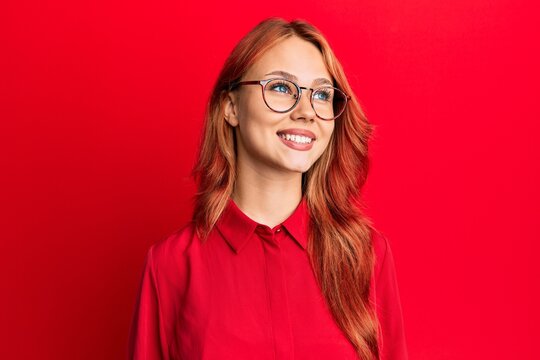 Young beautiful redhead woman wearing casual clothes and glasses over red background looking away to side with smile on face, natural expression. laughing confident.