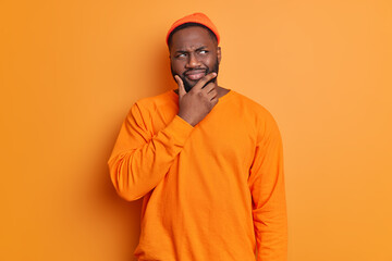 Studio shot of handsome bearded Afro American man holds chin looks thoughtfully aside thinks deeply about something wears hat and sweater poses against bright vivid orange background. Let me think