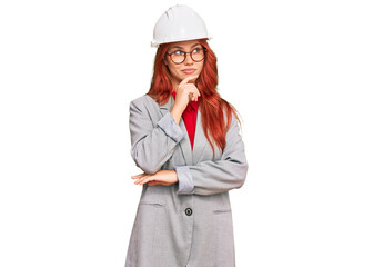 Young redhead woman wearing architect hardhat with hand on chin thinking about question, pensive expression. smiling with thoughtful face. doubt concept.