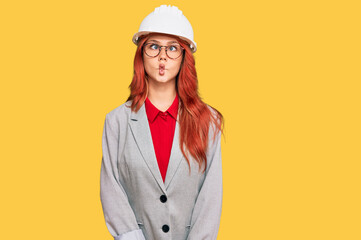 Young redhead woman wearing architect hardhat making fish face with lips, crazy and comical gesture. funny expression.