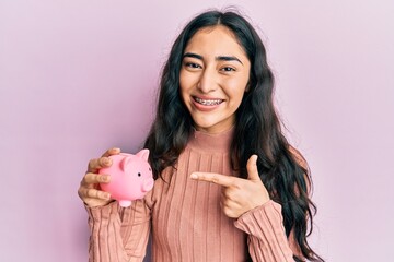 Hispanic teenager girl with dental braces holding piggy bank smiling happy pointing with hand and...