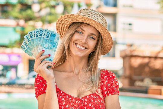 Young blonde tourist woman wearing summer style holding brazilian 100 real banknotes at the city.