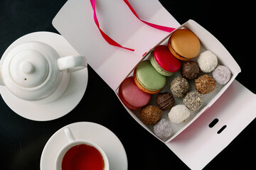 Composition with colorful macaroons, sweets truffle and custard dessert shu in paper box teapot and up of tea on dark background. Gift sets of sweets for Valentine's Day, February 14 and March 8.