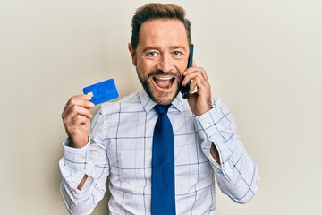 Middle age businessman talking on the smartphone holding credit card smiling and laughing hard out loud because funny crazy joke.