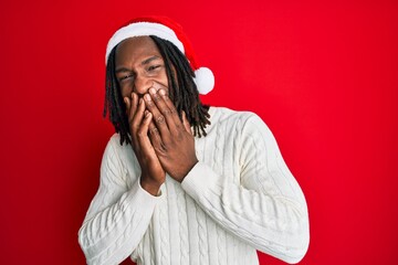 African american man with braids wearing christmas hat laughing and embarrassed giggle covering...