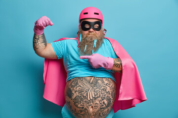 Serious bearded adult European man raises arm points at muscles dressed in superhero costume has...