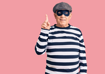 Senior handsome man wearing burglar mask and t-shirt pointing finger up with successful idea. exited and happy. number one.