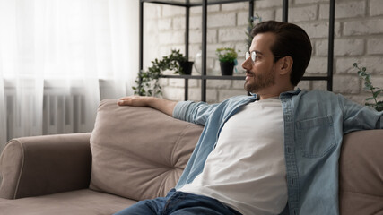 Close up dreamy calm young man wearing glasses looking to aside, sitting relaxing on cozy couch at home, thinking about new opportunities, dreaming or visualizing good future, lost in thoughts