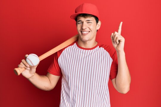 Handsome caucasian man playing baseball holding bat and ball surprised with an idea or question pointing finger with happy face, number one