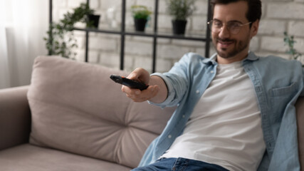 Close up smiling young man wearing glasses using remote tv controller, sitting on couch, relaxing, watching movie or show, switching channels, spending leisure time with television at home