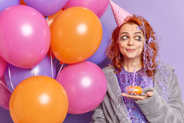 Fototapeta na wymiar People and holidays concept. Pleased thoughtful redhead young woman looks away wears soft dressing gown and party hat thinks what wish to make before blowing candle poses with colorful balloons