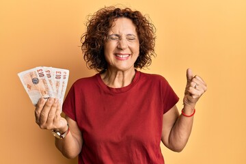 Beautiful middle age mature woman holding 10 united kingdom pounds banknotes screaming proud,...