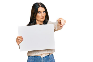 Middle age brunette woman holding blank empty banner pointing with finger to the camera and to you, confident gesture looking serious