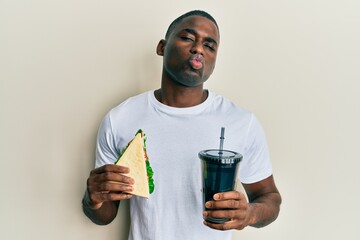 Young african american man eating sandwich and drinking soda looking at the camera blowing a kiss being lovely and sexy. love expression.