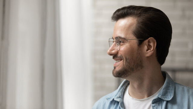 Close up side view smiling dreamy young man wearing glasses looking to aside, standing near window at home, businessman thinking about new opportunities, dreaming or visualizing good future