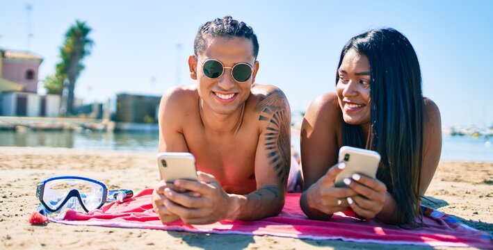 Young latin couple using smartphone lying on the sand at the beach.