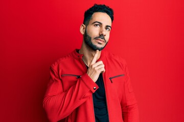 Young hispanic man wearing red leather jacket thinking concentrated about doubt with finger on chin and looking up wondering