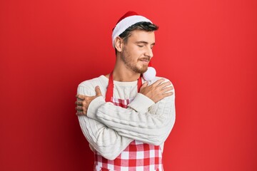 Handsome caucasian man wearing baker apron and christmas hat hugging oneself happy and positive, smiling confident. self love and self care