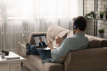 Obraz premium Rear view man wearing headphones using laptop, chatting with colleague online, discussing project strategy, making video call, businessman working at home, sitting on cozy couch in living room