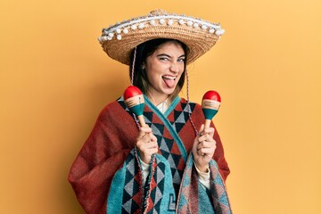 Young caucasian woman holding mexican hat using maracas sticking tongue out happy with funny...