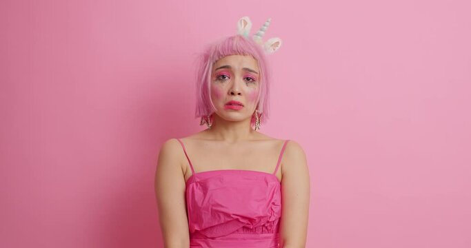 Discontent young Asian female model sobbing after crying has grief expression frustrated because of something wears rosy dress isolated over pink background. People negative emotions concept