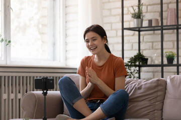 Overjoyed woman blogger shooting video on smartphone standing on tripod at home, smiling attractive female influencer recording vlog, chatting with friends online, engaged in online conference