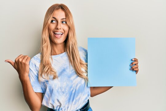 Beautiful blonde young woman holding blank empty blue banner pointing thumb up to the side smiling happy with open mouth