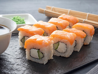 Sushi rolls philadelphia on a black textured plate stand on a gray background. wasabi ginger and sauce chopsticks. Rolls with salmon, cream cheese, avocado.
